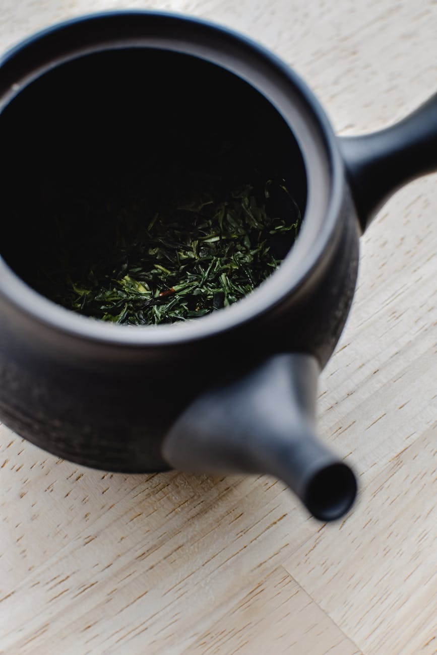 Steaming green sencha tea leaves unfurling in a white ceramic cup, releasing pale golden liquid against a minimalist background.