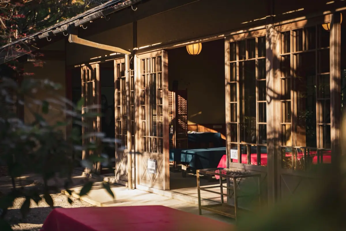 Traditional Japanese tea room showing low ceiling with exposed wooden beams, tatami flooring, tokonoma alcove, and small square window casting natural light.