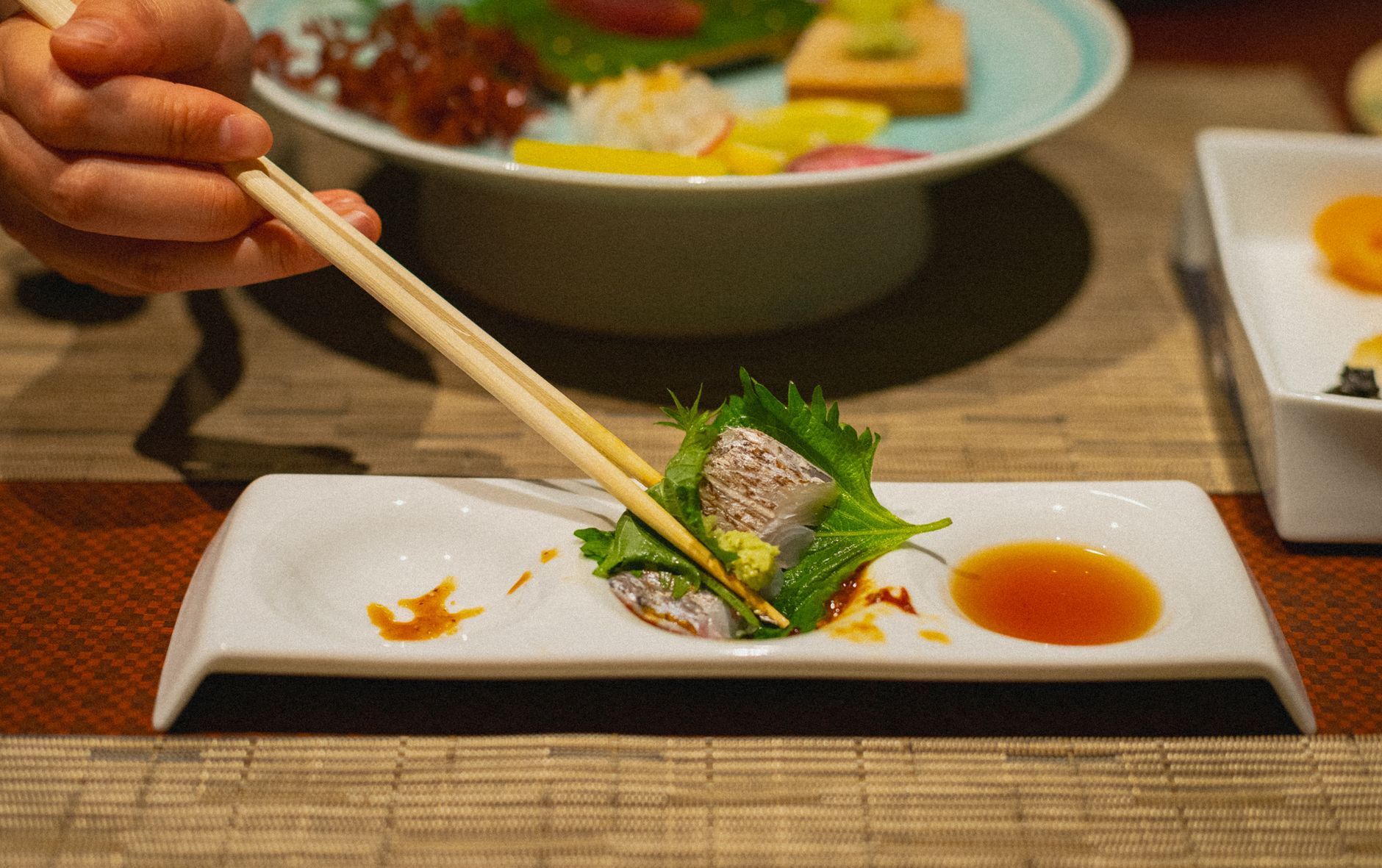 Hand holding lacquered chopsticks in proper tripod grip position over a ceramic bowl on a wooden table.