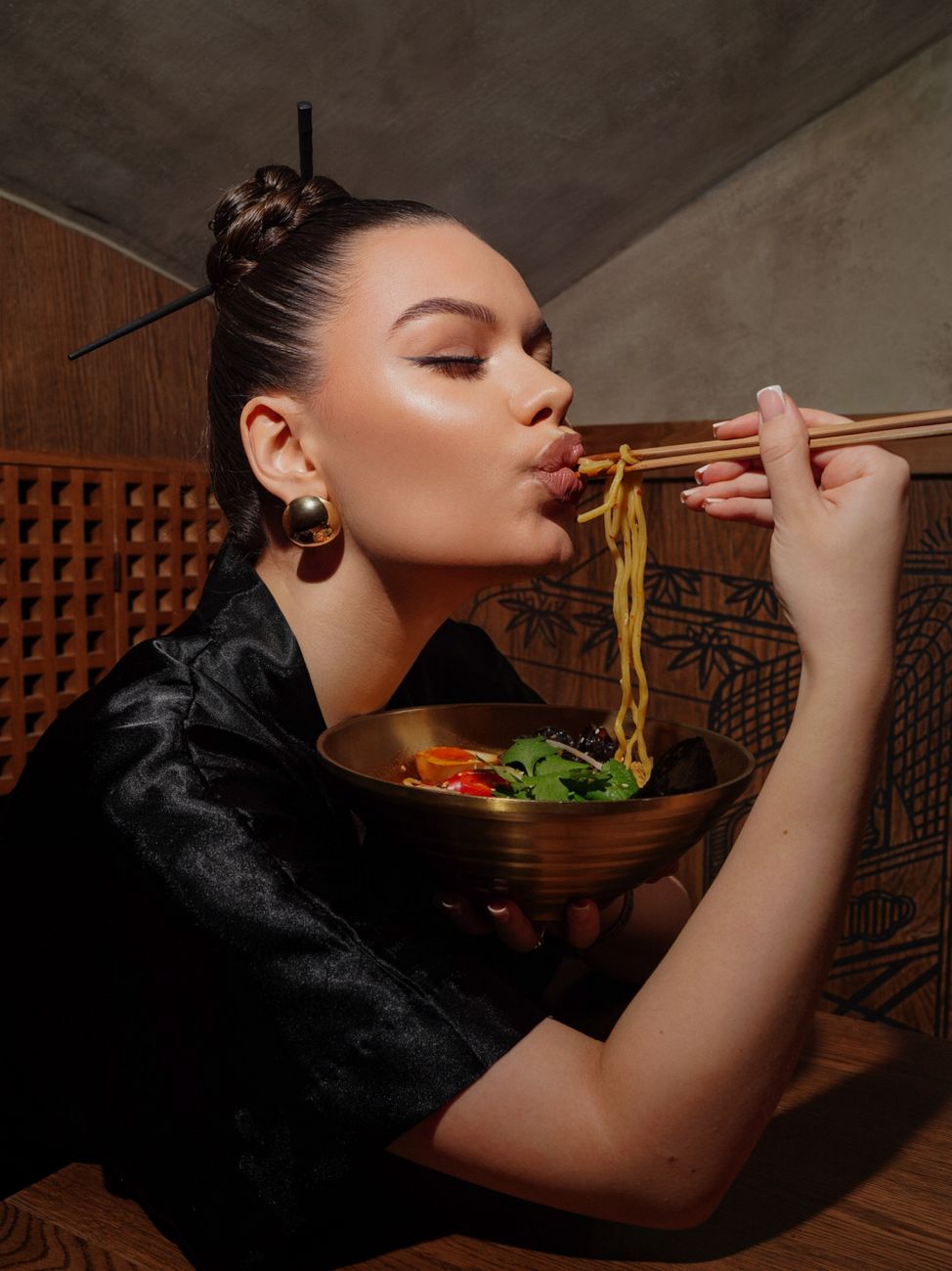 Hand holding lacquered chopsticks in proper tripod grip position over a ceramic bowl on a wooden table.