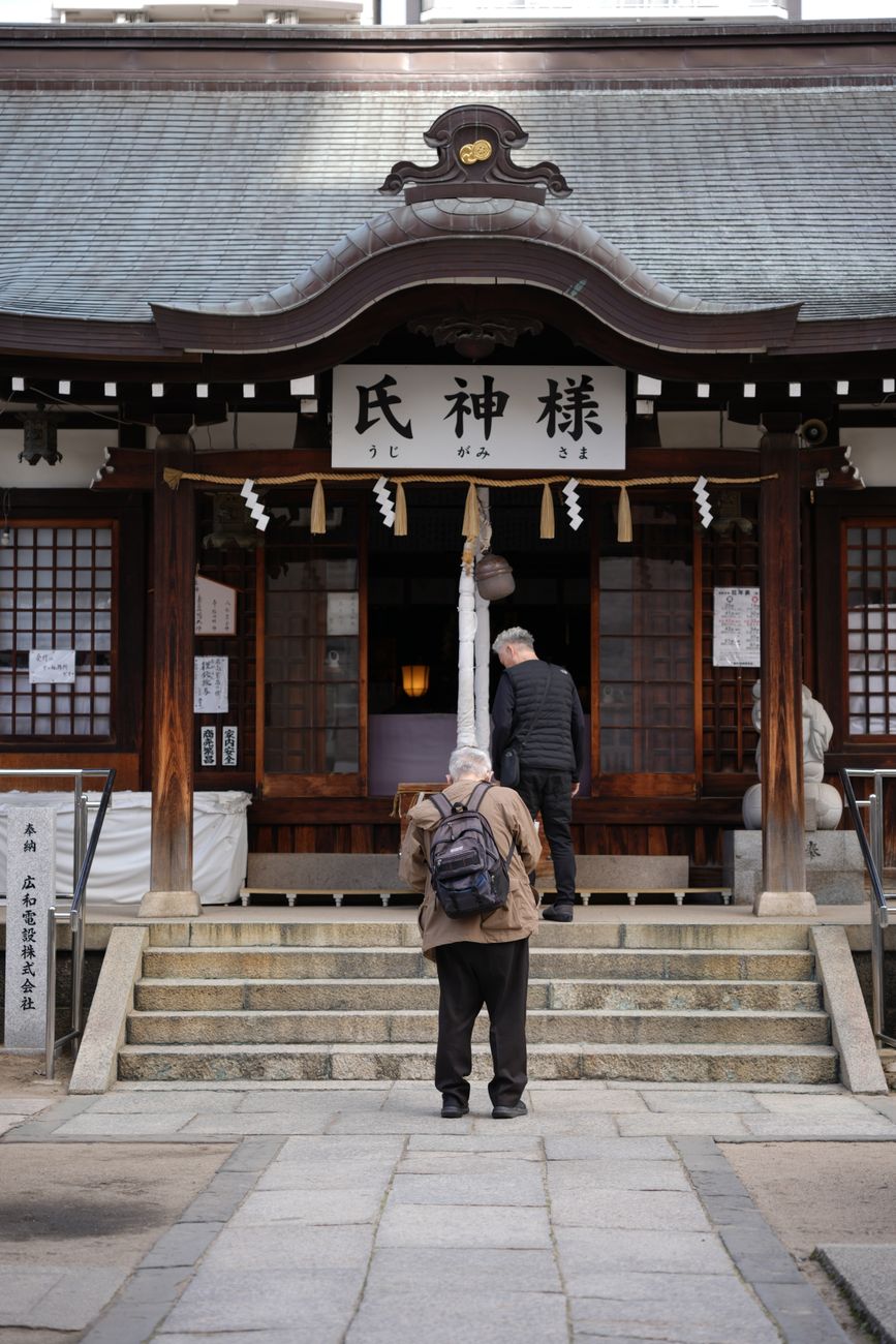 Visitor bowing at a wooden shrine torii gate with stone lanterns lining the gravel path in morning light.