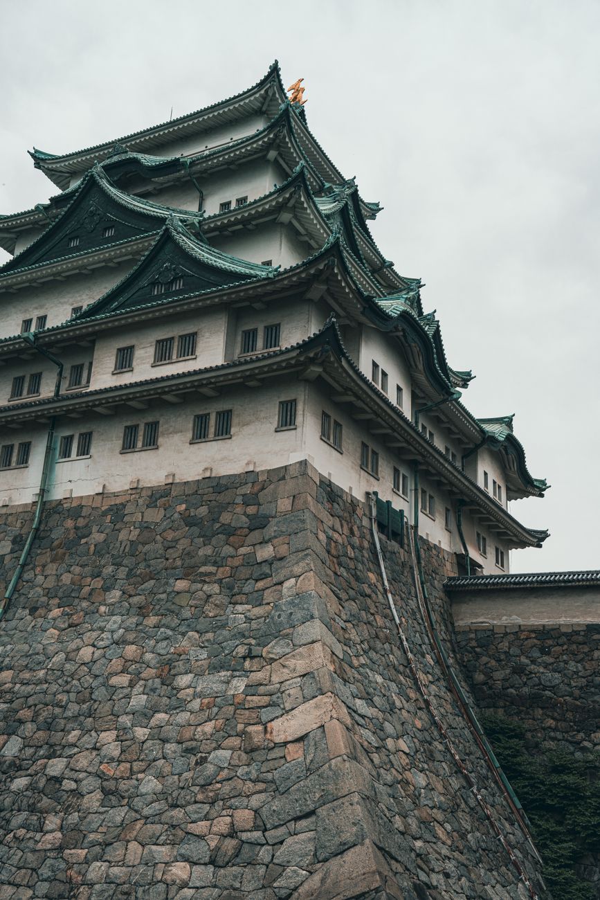 Massive curved stone walls rise in precise tiers beneath a multi-story wooden castle tower with white plaster and dark timber.