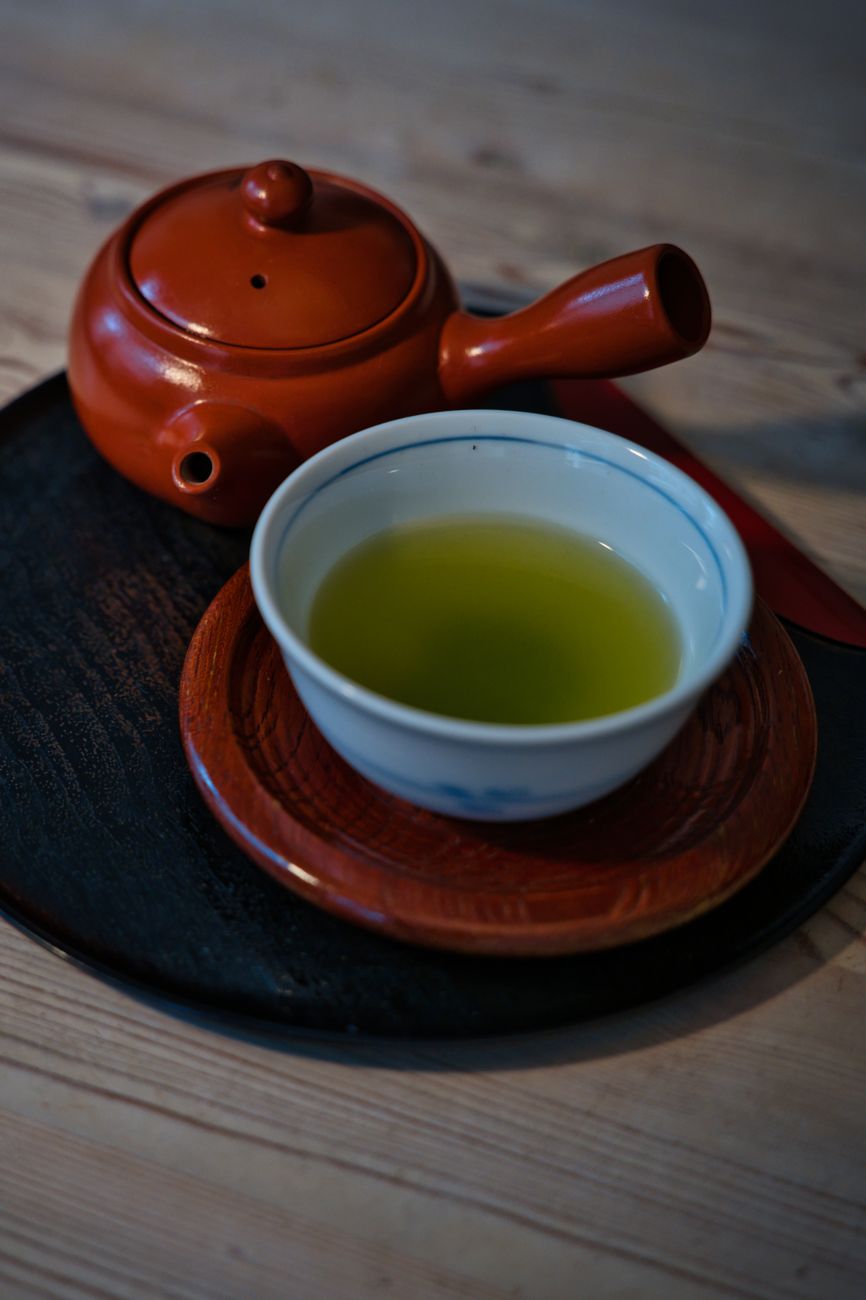 Vibrant green matcha powder in a ceramic bowl beside fresh sencha tea leaves on a bamboo mat.