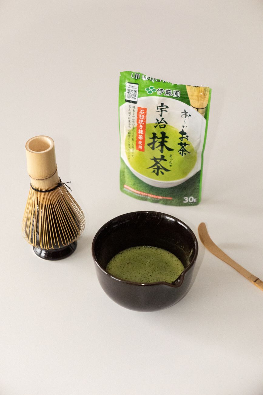 Traditional Japanese tea ceremony host whisking vibrant green matcha powder in a black ceramic bowl using bamboo whisk.