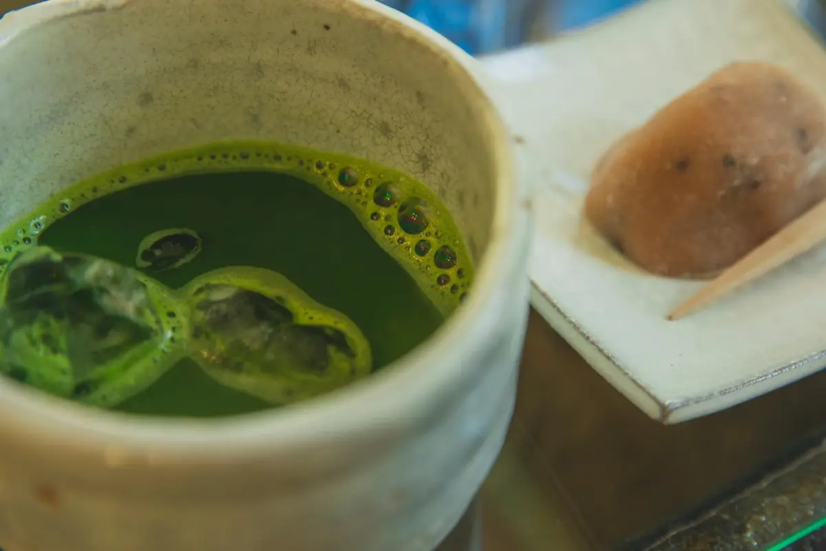 Side-by-side comparison showing bright green matcha powder in a bowl next to loose sencha tea leaves on a bamboo mat.