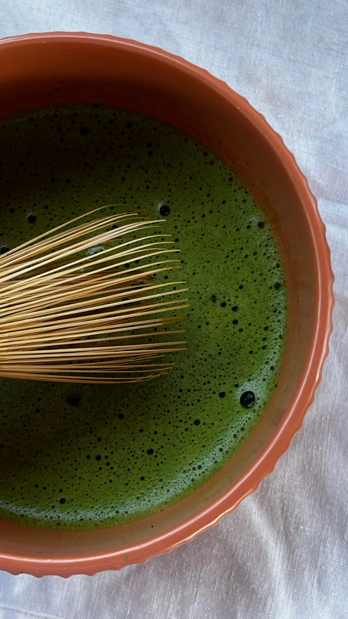 A bamboo whisk whisking thin, bright green usucha matcha in a ceramic tea bowl, creating a frothy layer on the surface.