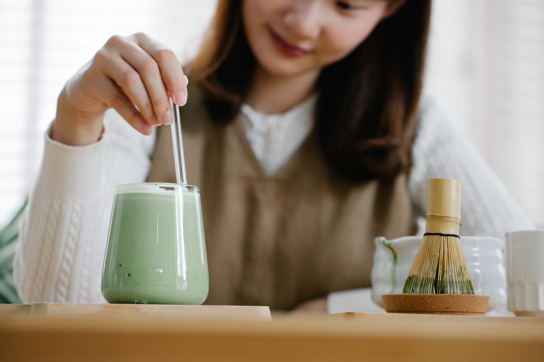 Bamboo whisk whisking bright green matcha powder in a ceramic bowl with steam gently rising from hot water.