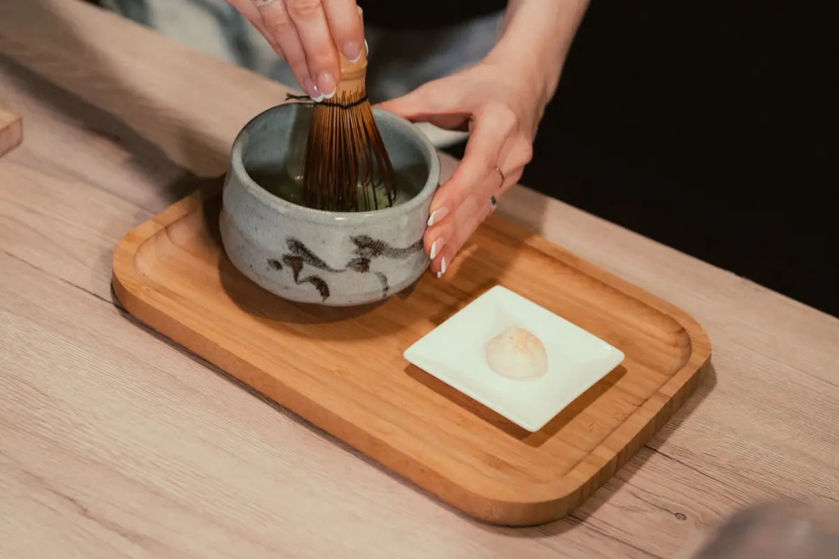 Fine mesh strainer positioned above a ceramic bowl, filtering bright green matcha powder to remove clumps before whisking.