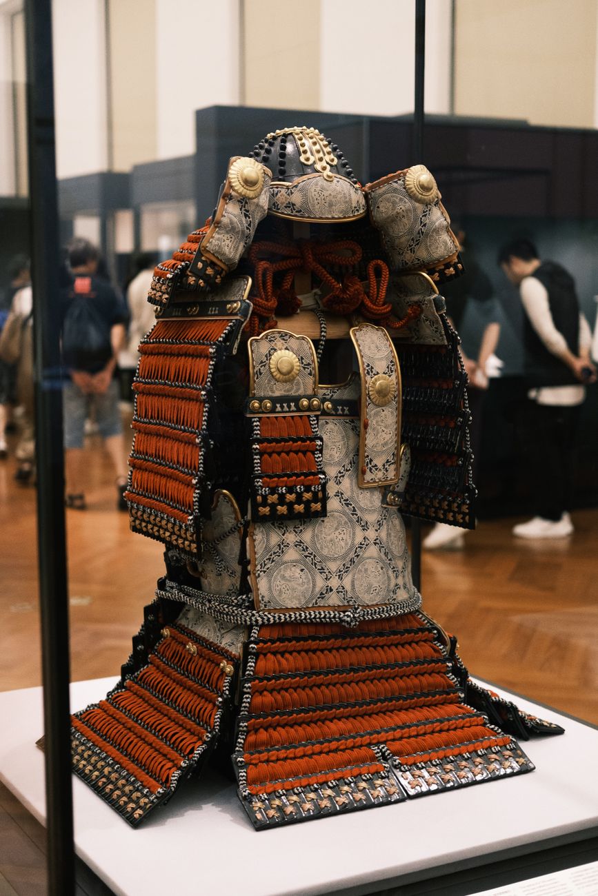 Samurai armor displayed in a traditional Japanese museum with calligraphy scrolls depicting the seven virtues of bushido on the wall behind.