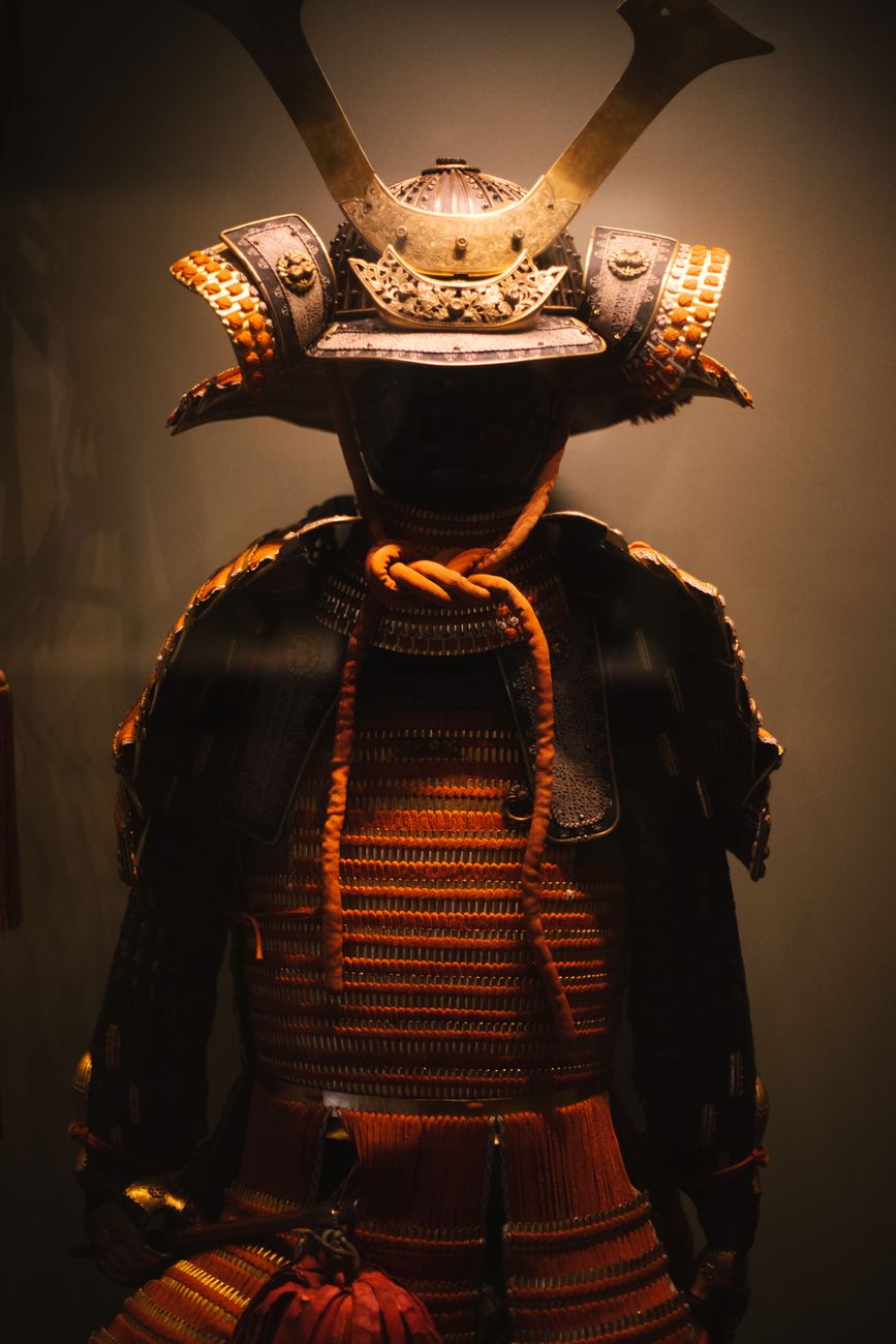 Complete traditional samurai armor displayed on a wooden stand, showing layered iron plates, ornate helmet with crescent crest, and protective shoulder guards.
