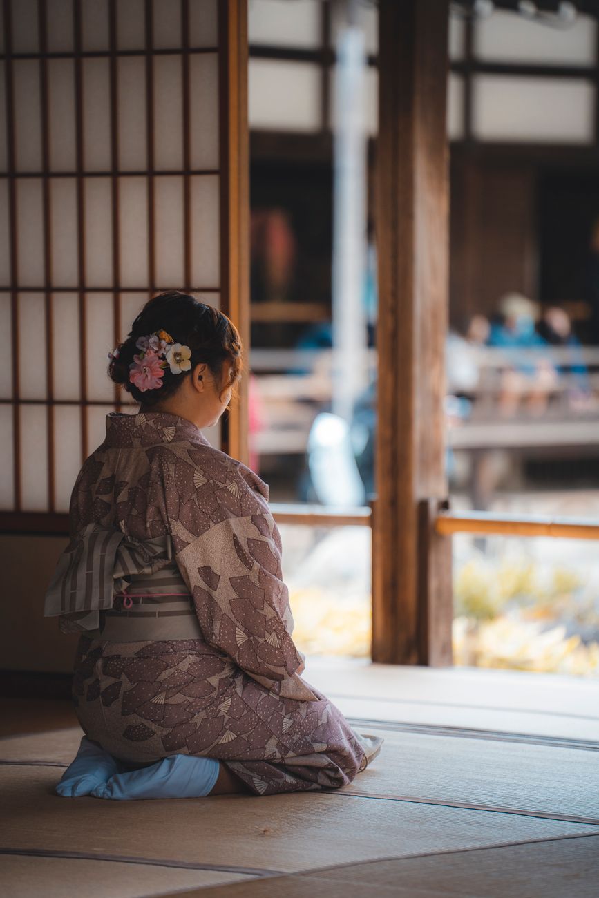 Person in traditional meditation posture sitting on zafu cushion in minimalist temple room with natural morning light filtering through shoji screens.