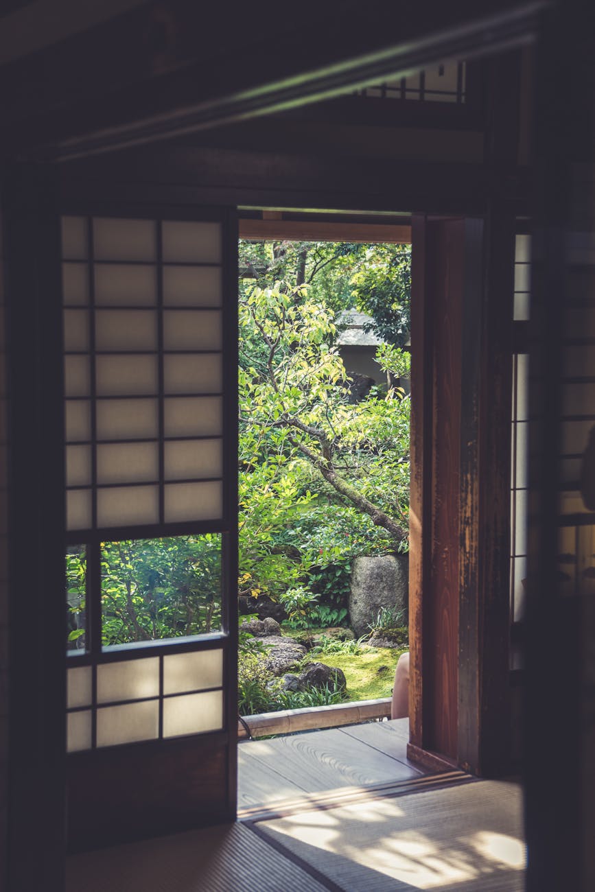 Person writing in a journal at a wooden desk near a window, surrounded by tea, plants, and morning sunlight.