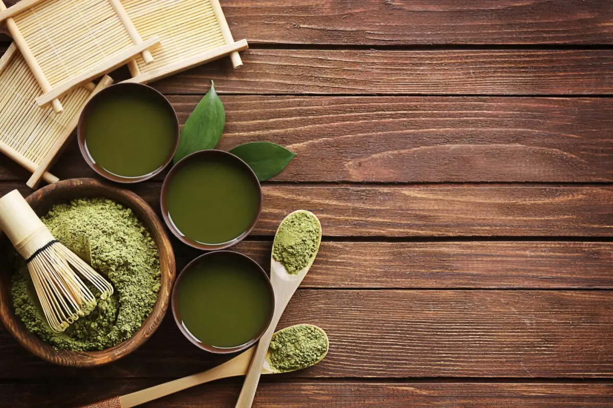 Vibrant green matcha powder in a ceramic bowl beside a bamboo whisk on a wooden table with natural light.