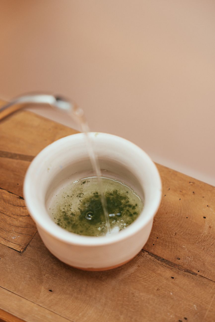 Vibrant green matcha powder in a ceramic bowl beside a bamboo whisk on a wooden table with natural light.