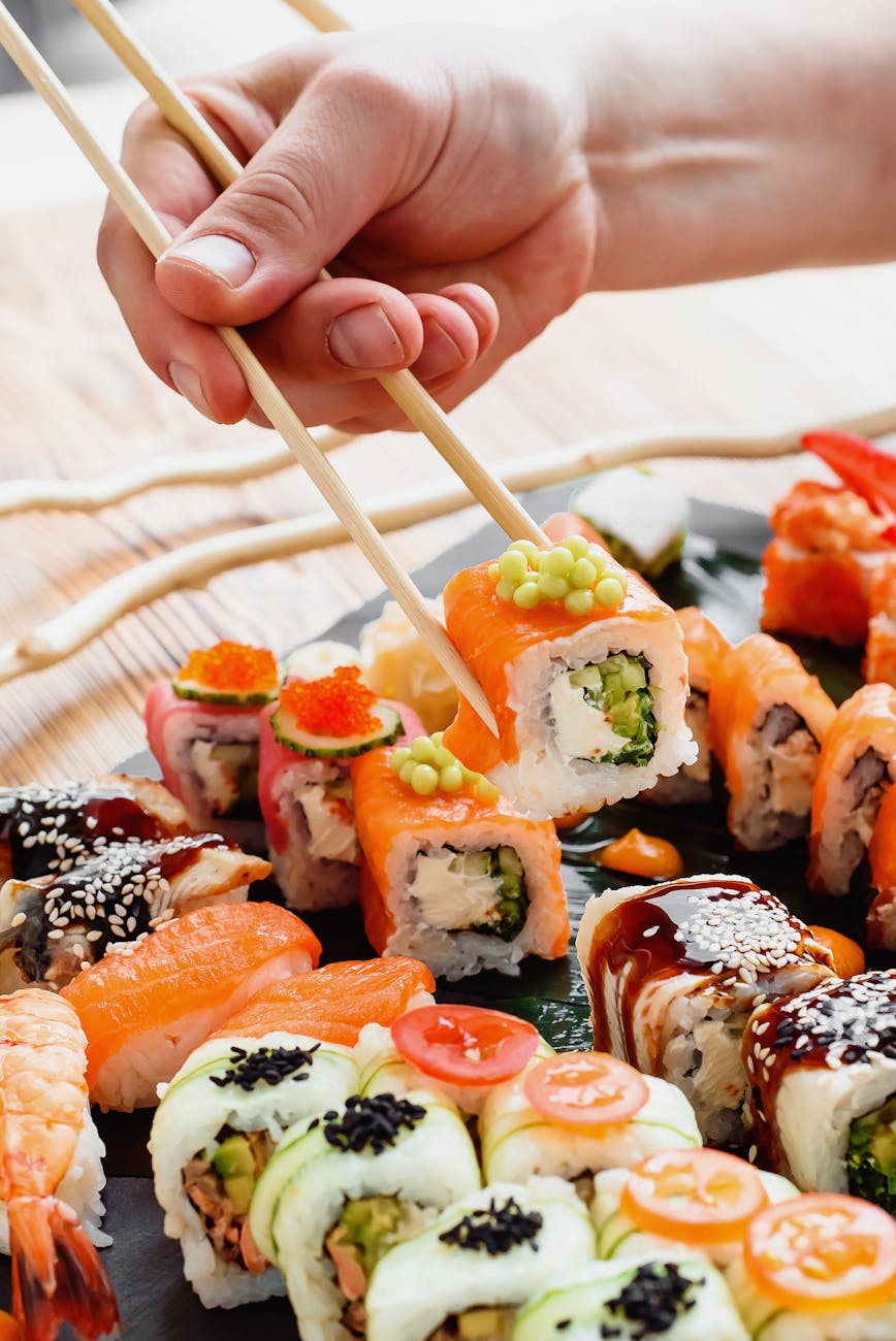 Hands holding various Japanese chopsticks made from wood, lacquer, and bamboo arranged on a traditional textile background.