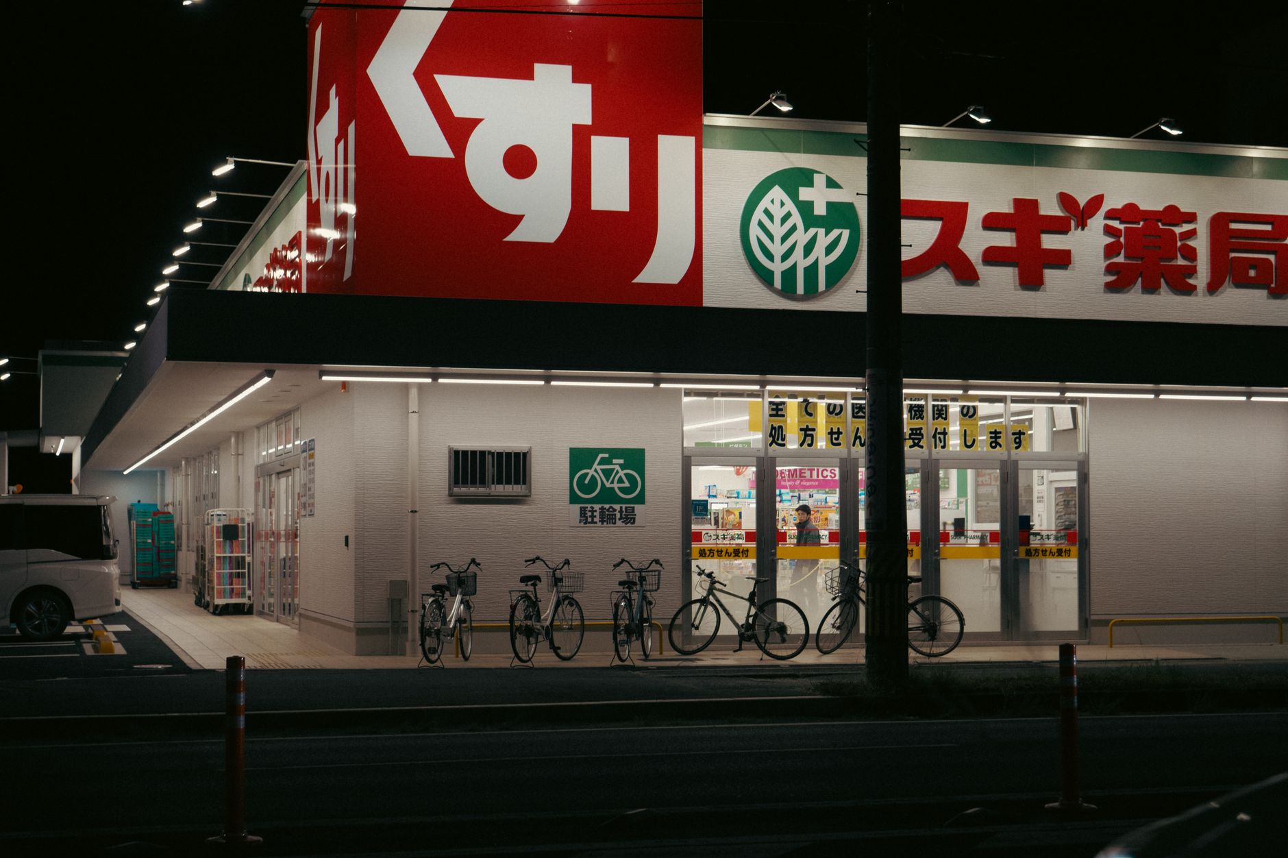 Illuminated Japanese convenience store exterior at night with bright signage, vending machines, and customers visible through glass windows.