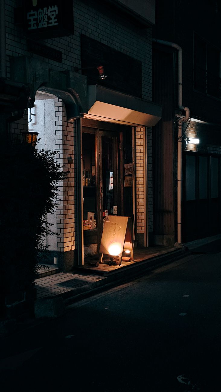 Illuminated Japanese convenience store exterior at night with bright signage, vending machines, and customers visible through glass windows.