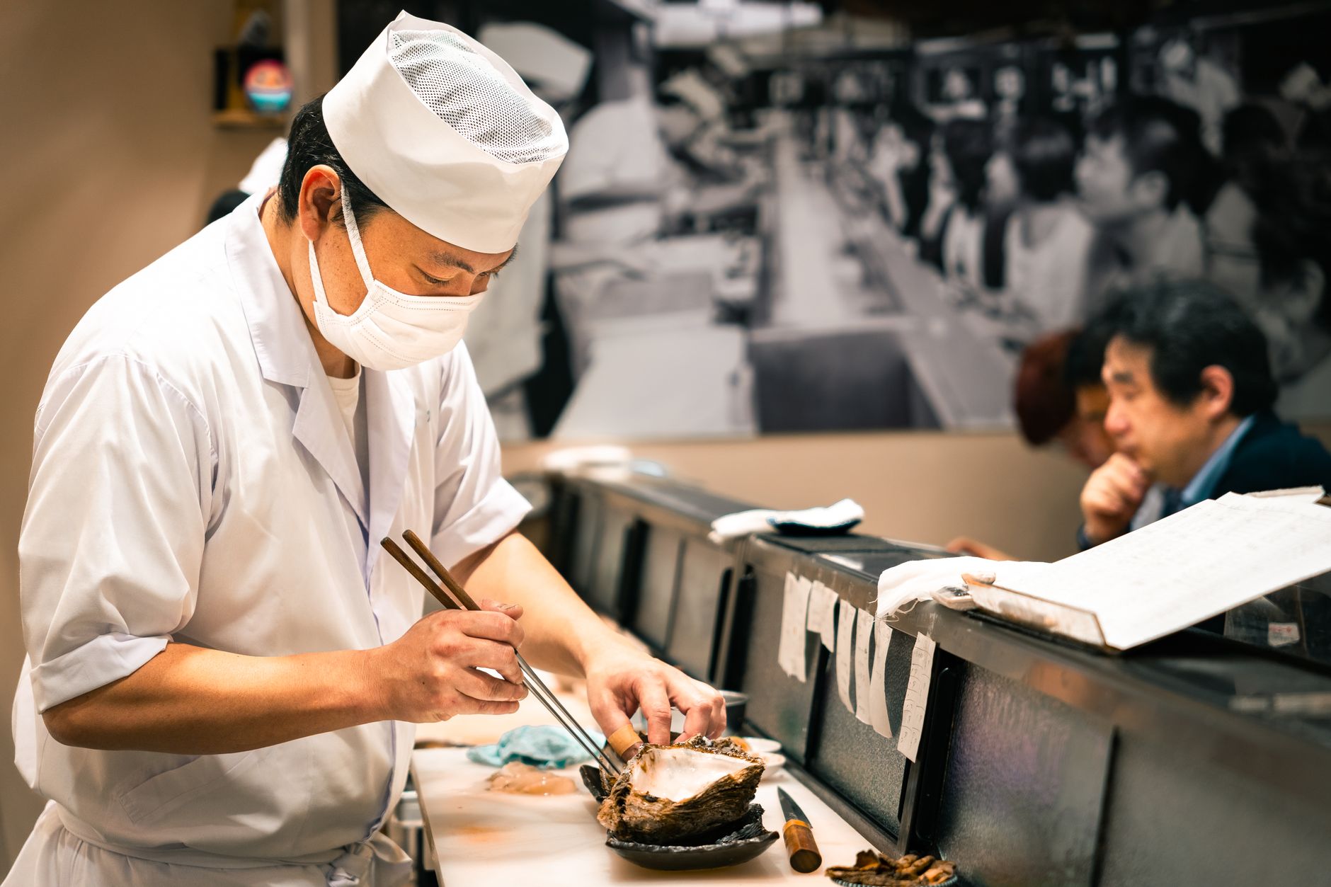 Japanese restaurant server bowing politely while presenting a wooden tray to a customer at a traditional counter seat.