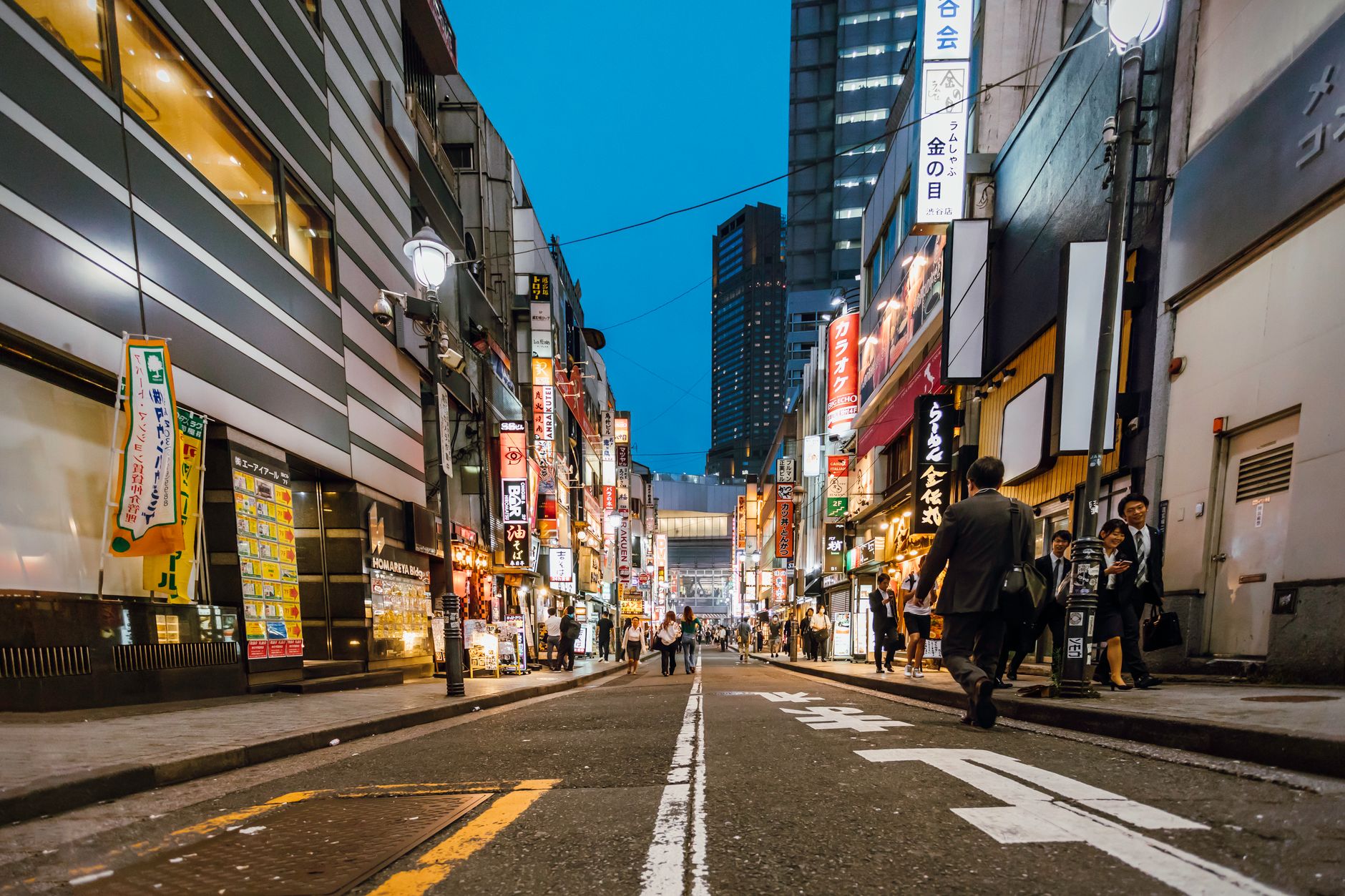 People walking on a busy Tokyo street wearing white surgical face masks during daytime with storefronts in background.