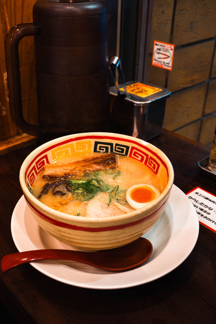 Person using chopsticks to lift steaming ramen noodles from a ceramic bowl, creating visible steam in a traditional Japanese setting.