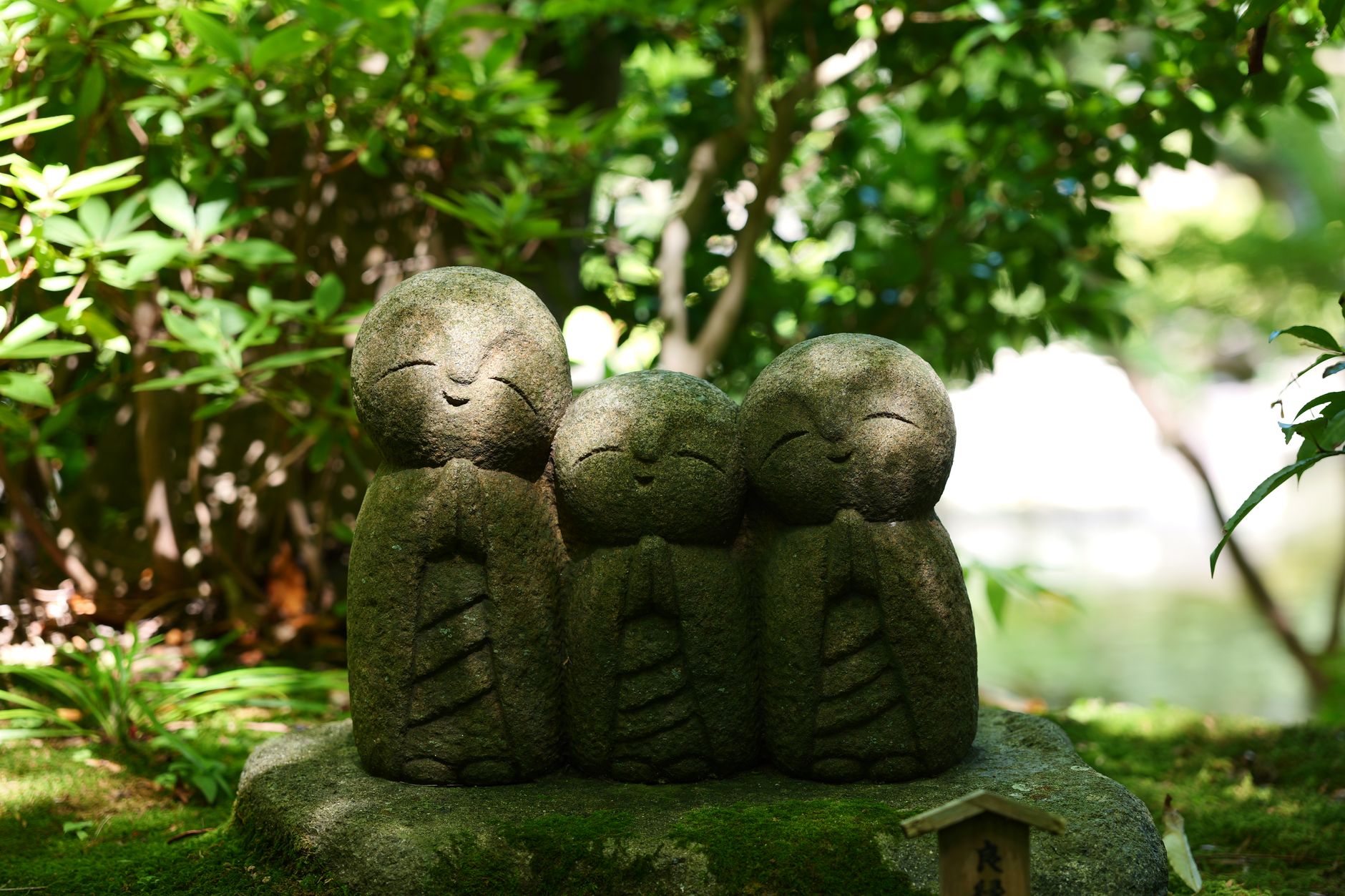 Small stone Jizo statue with red bib standing beside a rural Japanese roadside, covered in green moss and weathered by time.