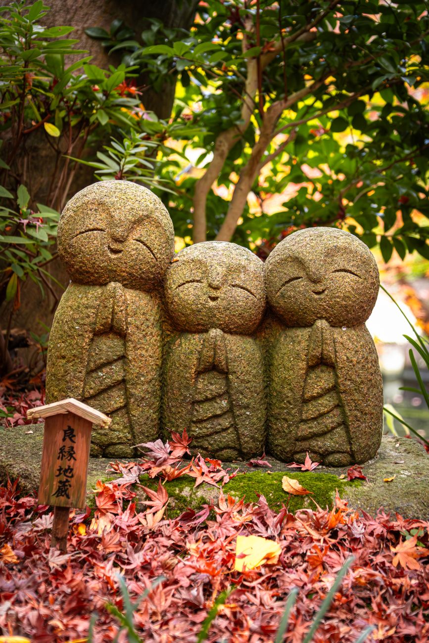 Small stone Jizo statue with red bib standing beside a rural Japanese roadside, covered in green moss and weathered by time.