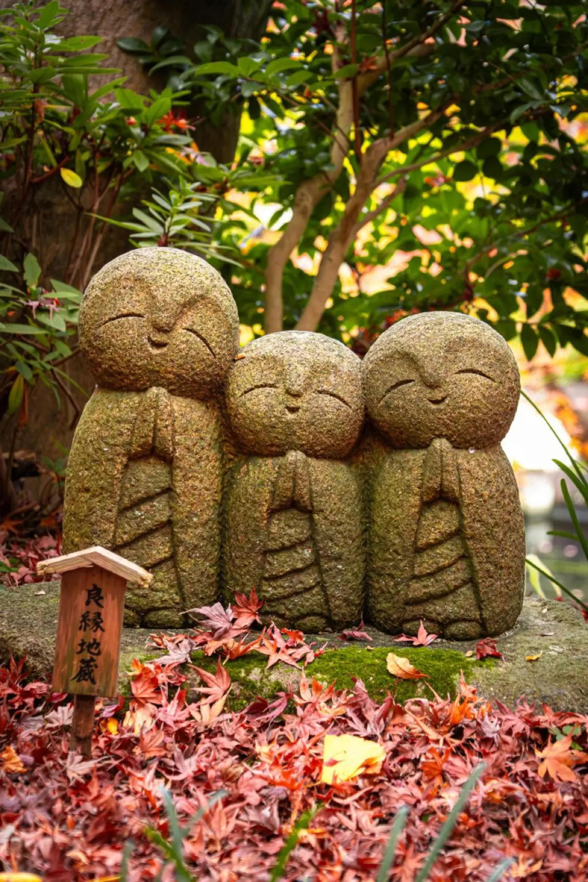 Small stone Jizo statue with red bib standing beside a rural Japanese roadside, covered in green moss and weathered by time.