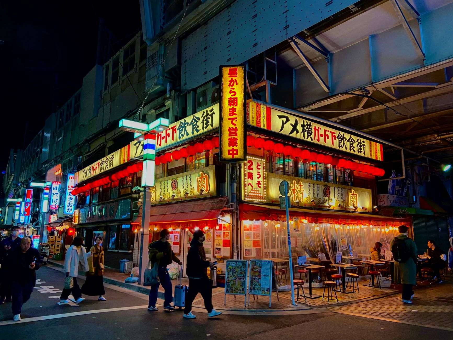 Colorful food stalls line a crowded Japanese festival street at night, with paper lanterns glowing and visitors browsing takoyaki and yakisoba vendors.