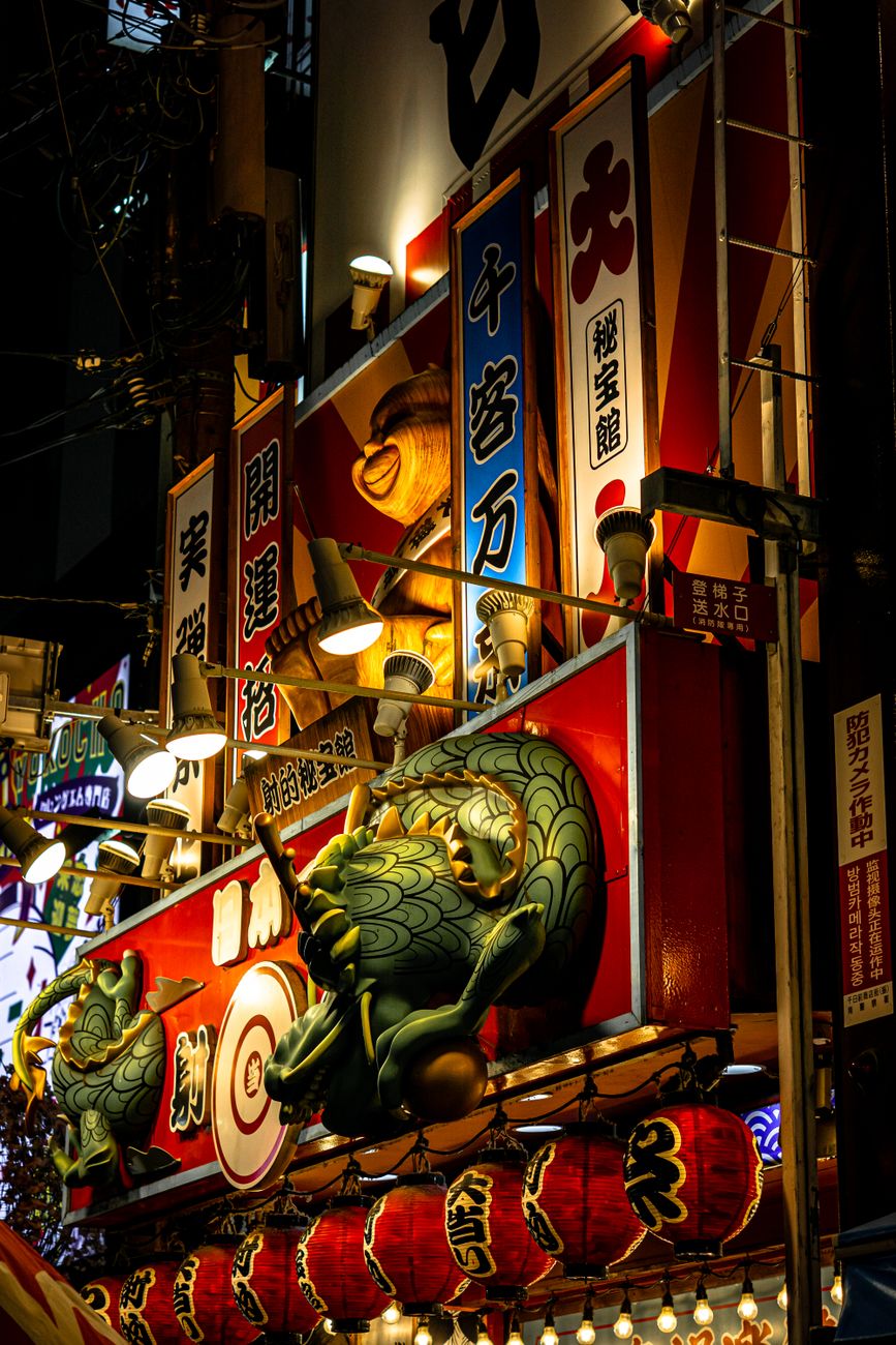 Colorful food stalls line a crowded Japanese festival street at night, with paper lanterns glowing and visitors browsing takoyaki and yakisoba vendors.