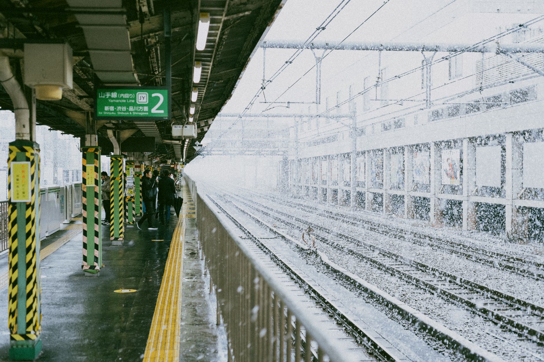 Passengers waiting on a Japanese train platform as a melody plays from overhead speakers before the train departs.