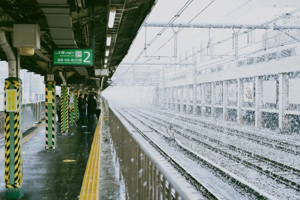 Passengers waiting on a Japanese train platform as a melody plays from overhead speakers before the train departs.