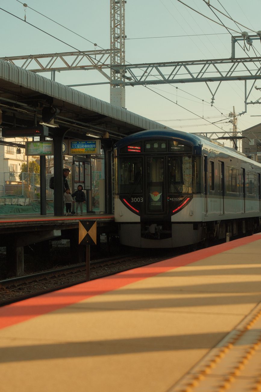 Passengers waiting on a Japanese train platform as a melody plays from overhead speakers before the train departs.