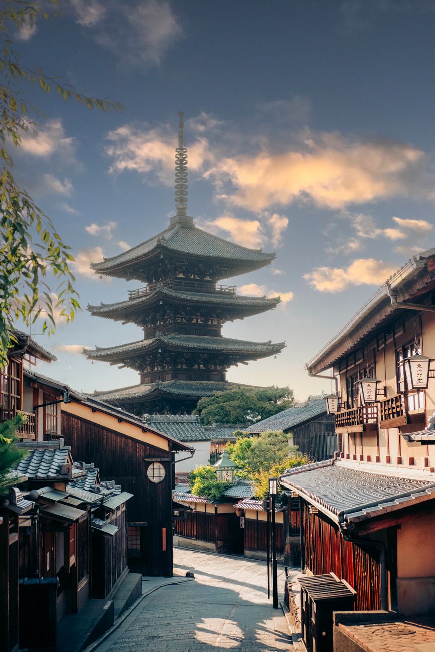 Traditional wooden pagoda rising above stone pathway lined with red torii gates in historic Kyoto temple district at sunset.