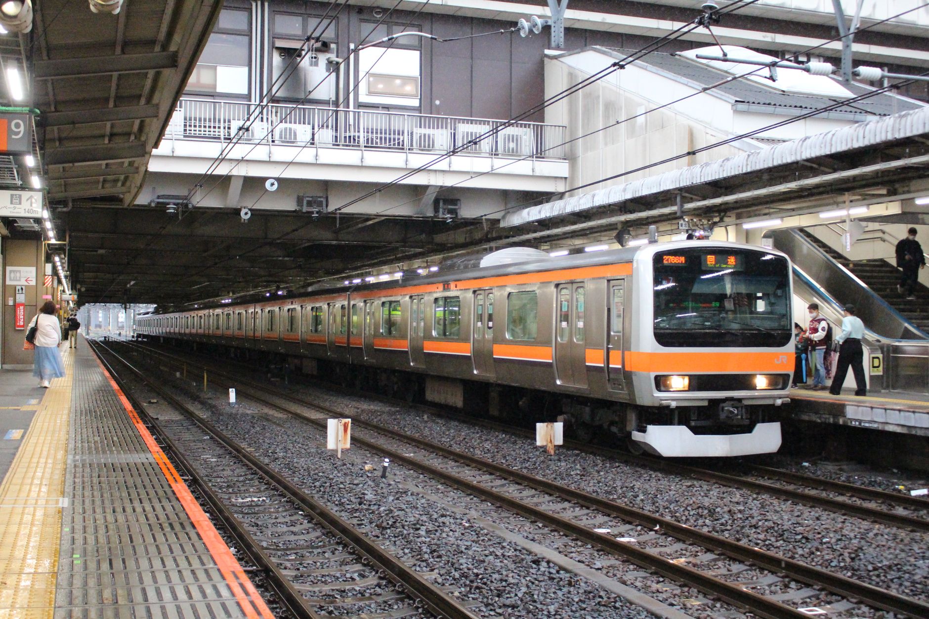 Japanese train conductor in uniform performing pointing and calling gesture toward departure board on railway platform.