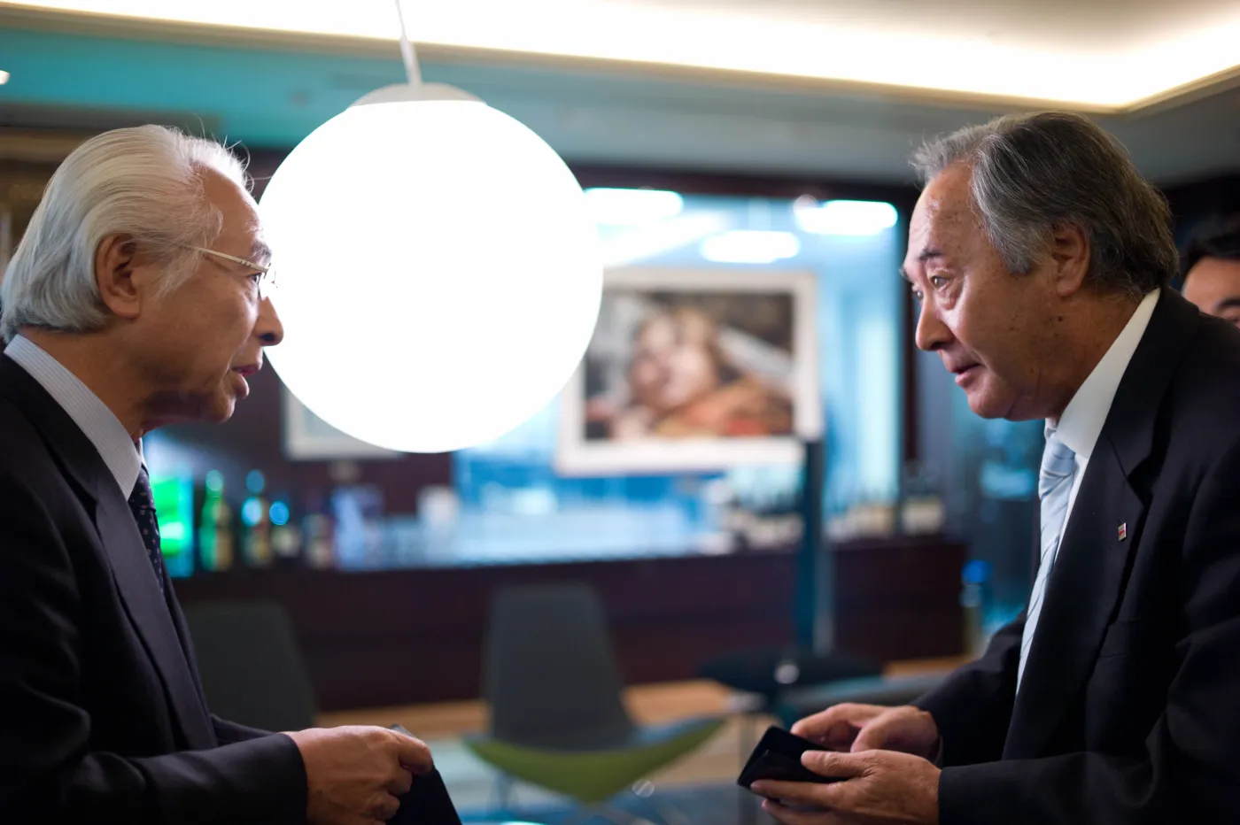 Two Japanese businesspeople in suits exchange business cards with both hands while bowing slightly in a modern office setting.