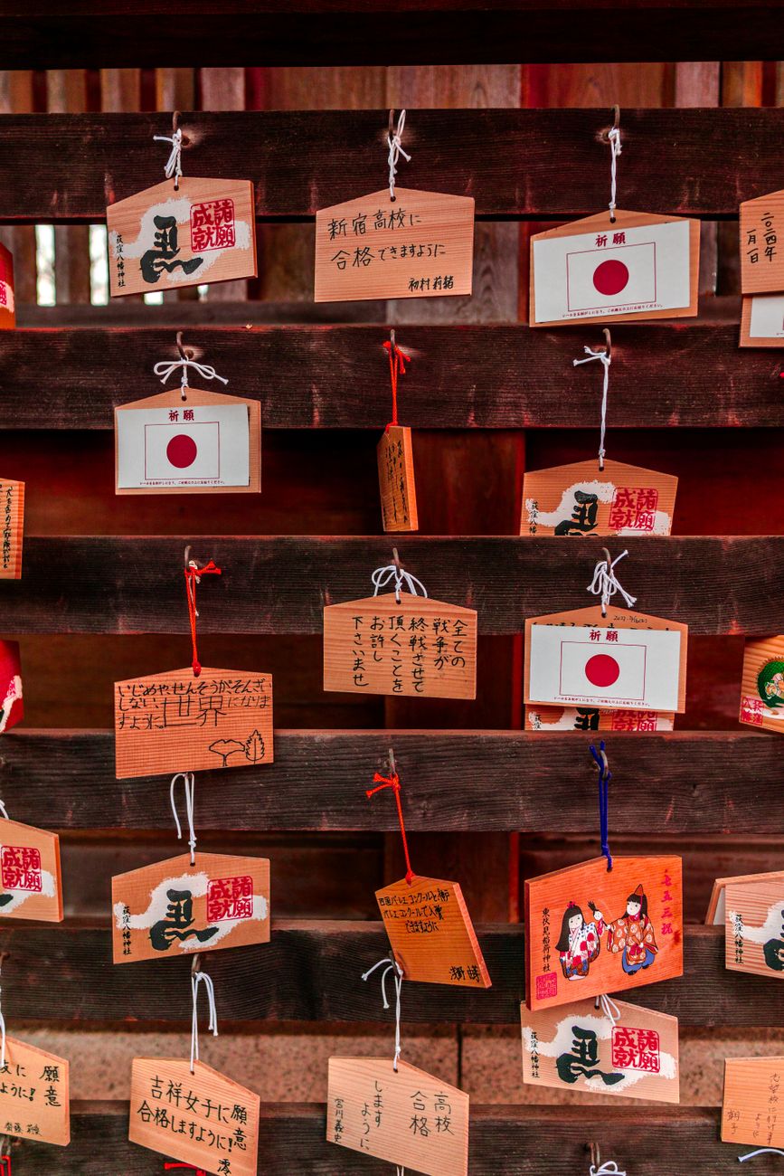 Small fabric omamori charm pouches in red, white, and gold hanging from wooden display at Japanese shrine.