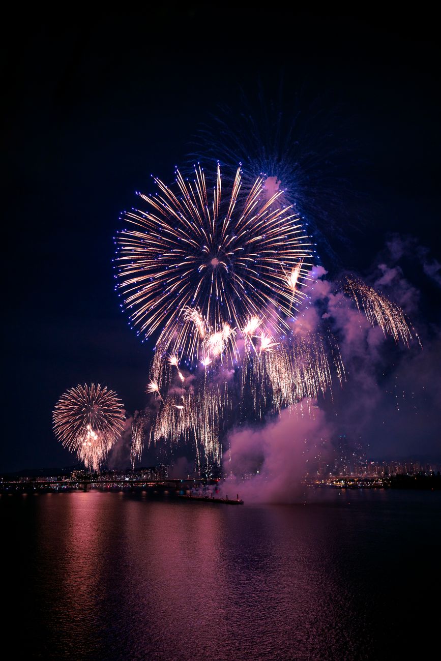 Colorful fireworks bursting over a Japanese riverside at dusk as crowds in yukata gather on blankets to watch the summer display.