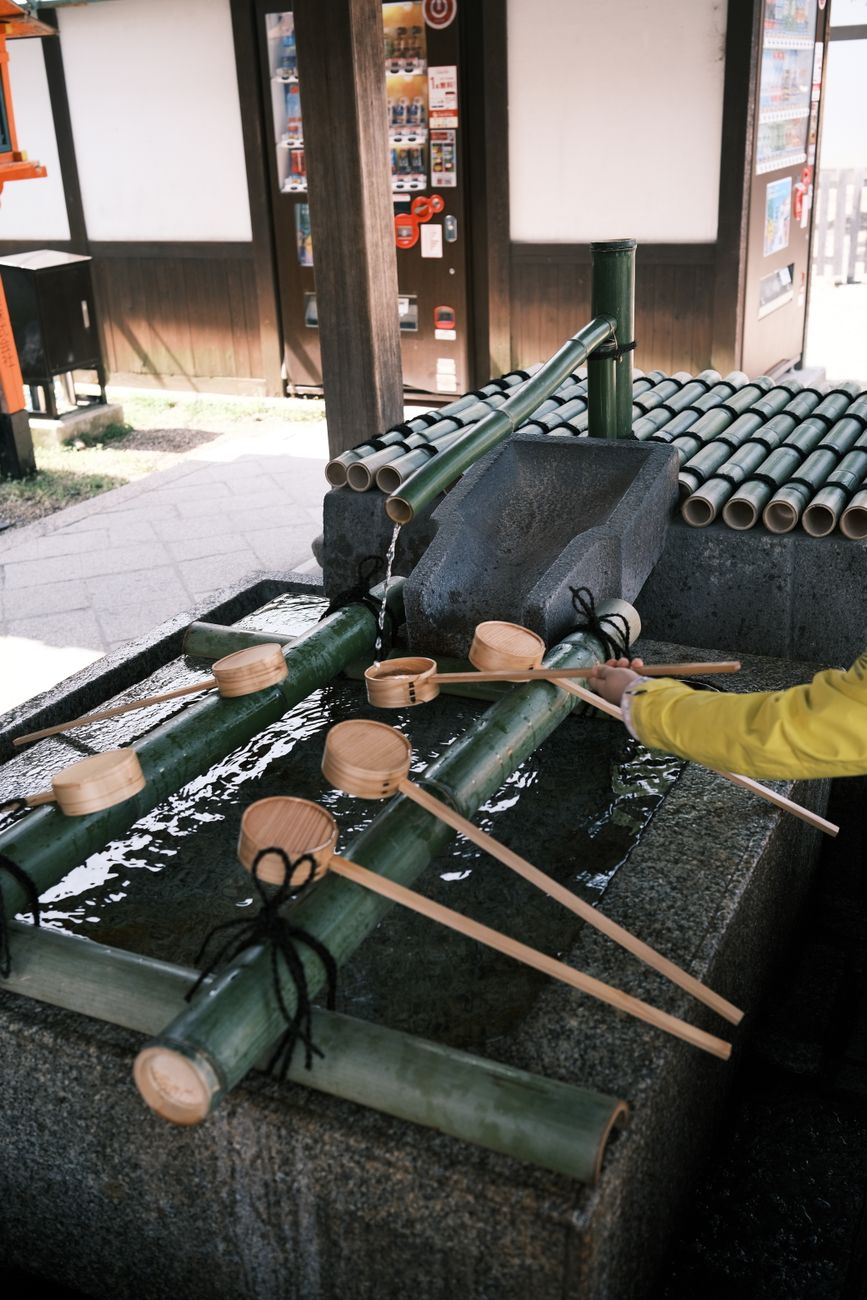 Visitors using long-handled ladles to rinse hands at a traditional stone water basin outside a Japanese shrine entrance.