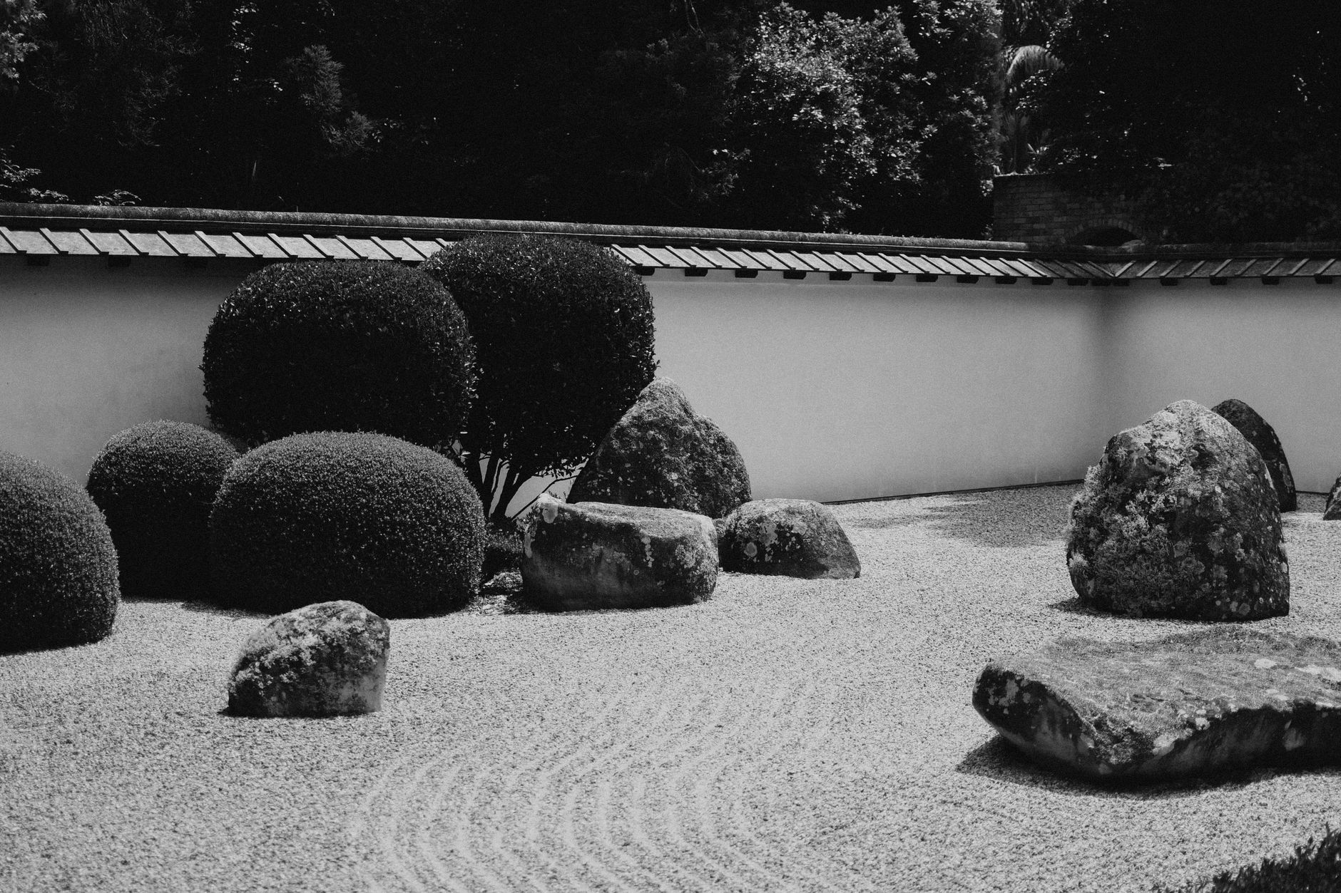 Monk in traditional robes carefully raking concentric circular patterns into white gravel around moss-covered stones in a Japanese zen garden.
