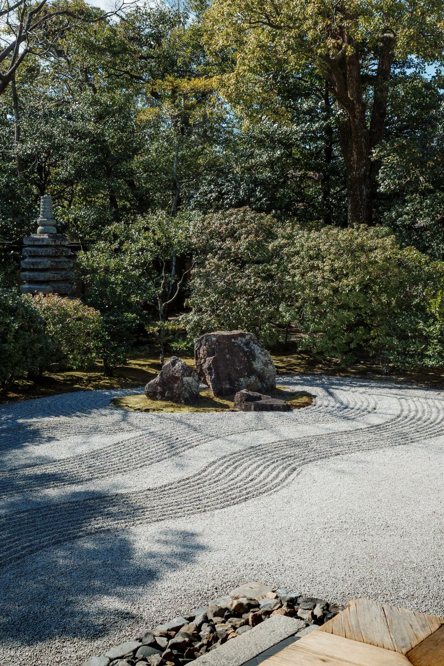 Monk in traditional robes carefully raking concentric circular patterns into white gravel around moss-covered stones in a Japanese zen garden.