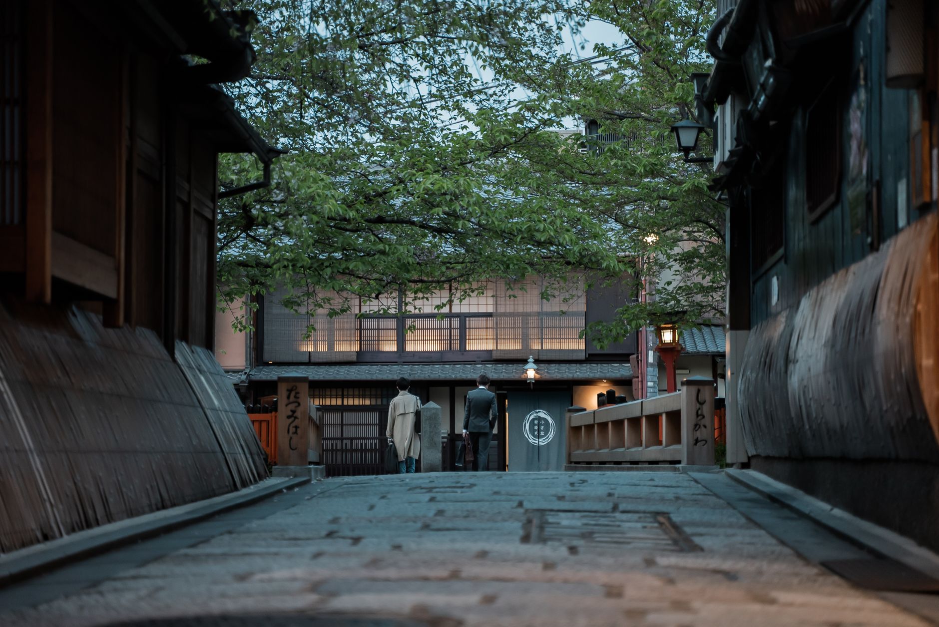 Traditional wooden machiya townhouses line a narrow stone-paved street in Gion at dusk with soft lantern light glowing.