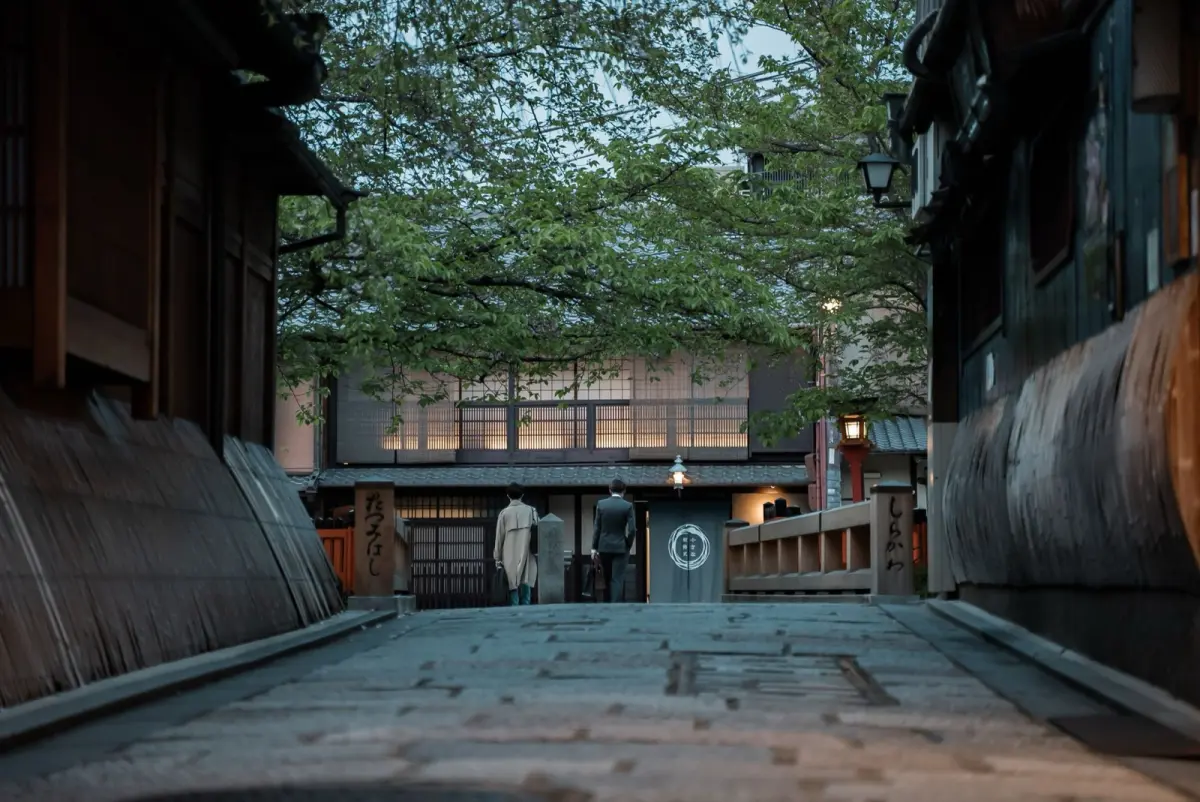 A maiko in kimono walking through a lantern-lit street in a historic Kyoto geisha district at dusk