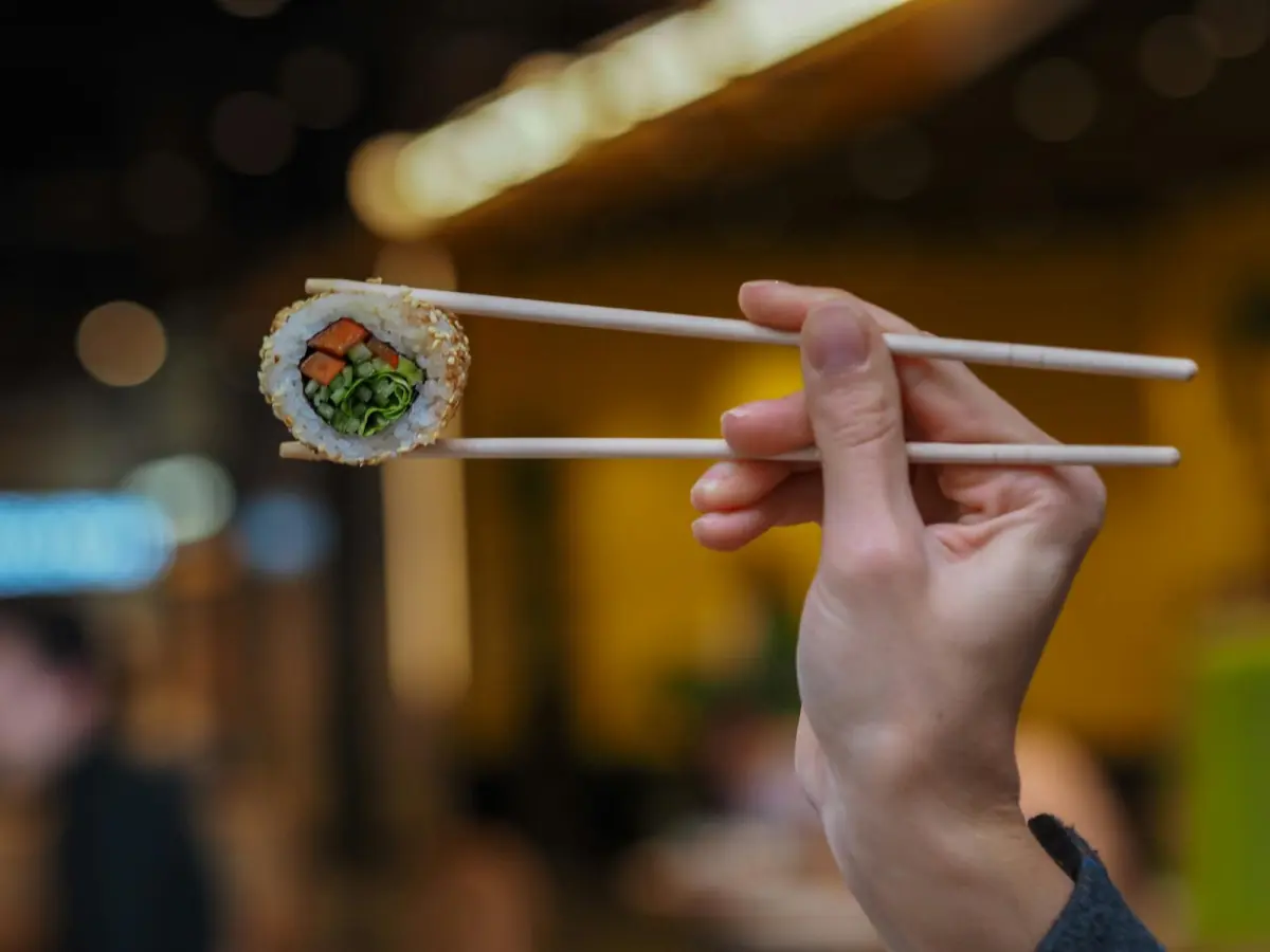 Hands demonstrating proper chopstick grip with thumb, index, and middle fingers positioned to hold two wooden chopsticks correctly.