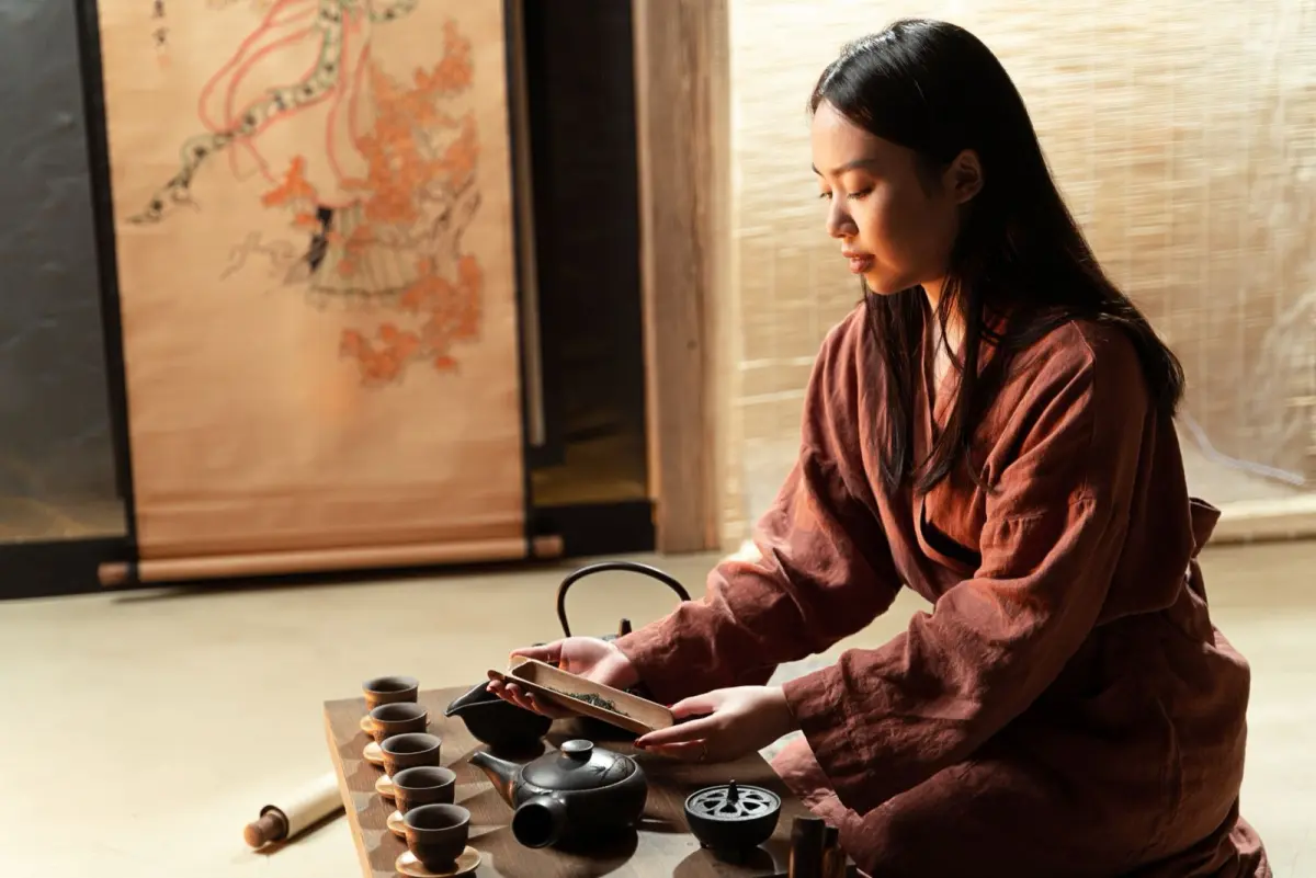 Elderly tea master in kimono performs precise hand movements while whisking matcha in black ceramic bowl during traditional ceremony.