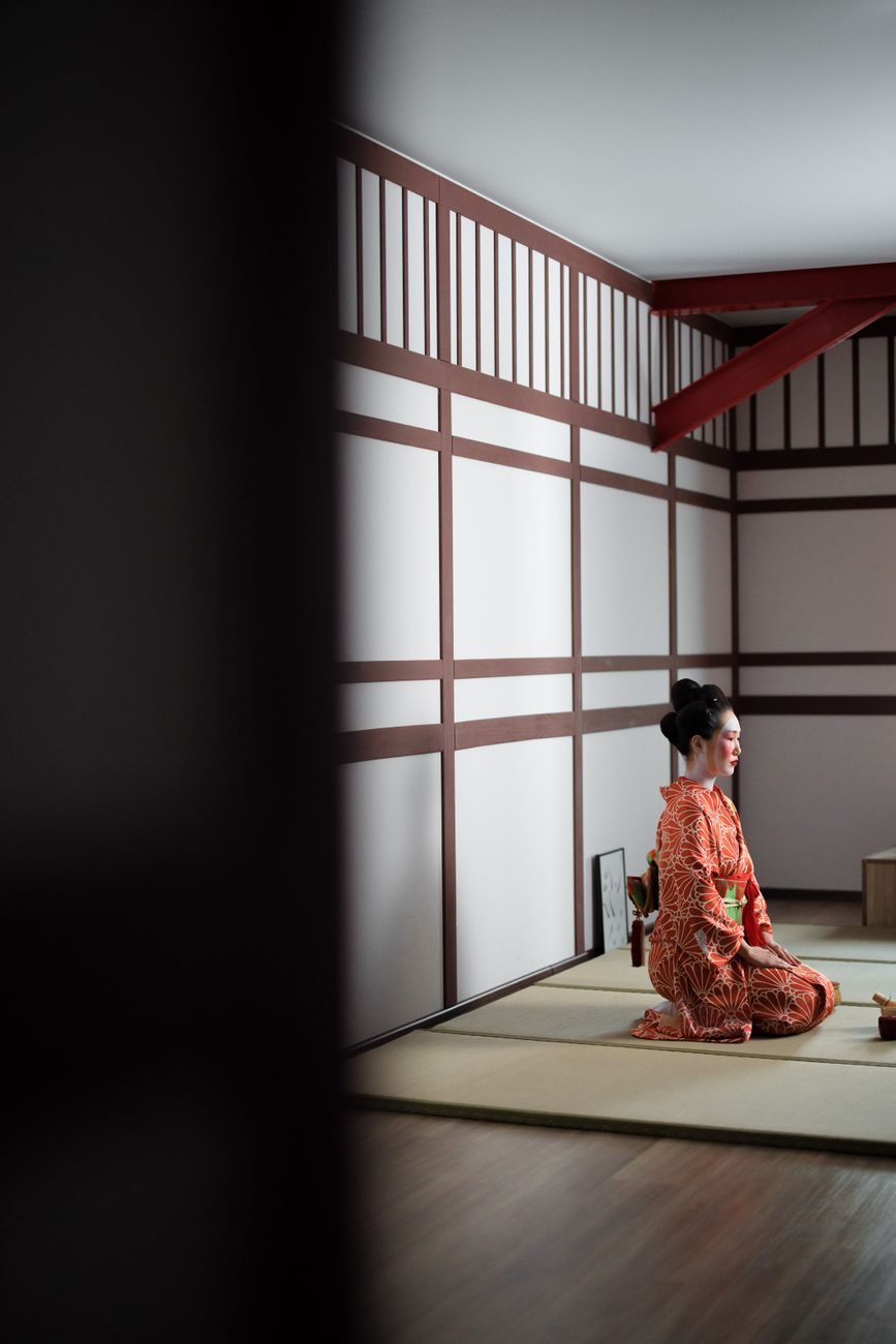 Traditional Japanese tea ceremony scene showing a host in kimono whisking matcha in a ceramic bowl on tatami mats.
