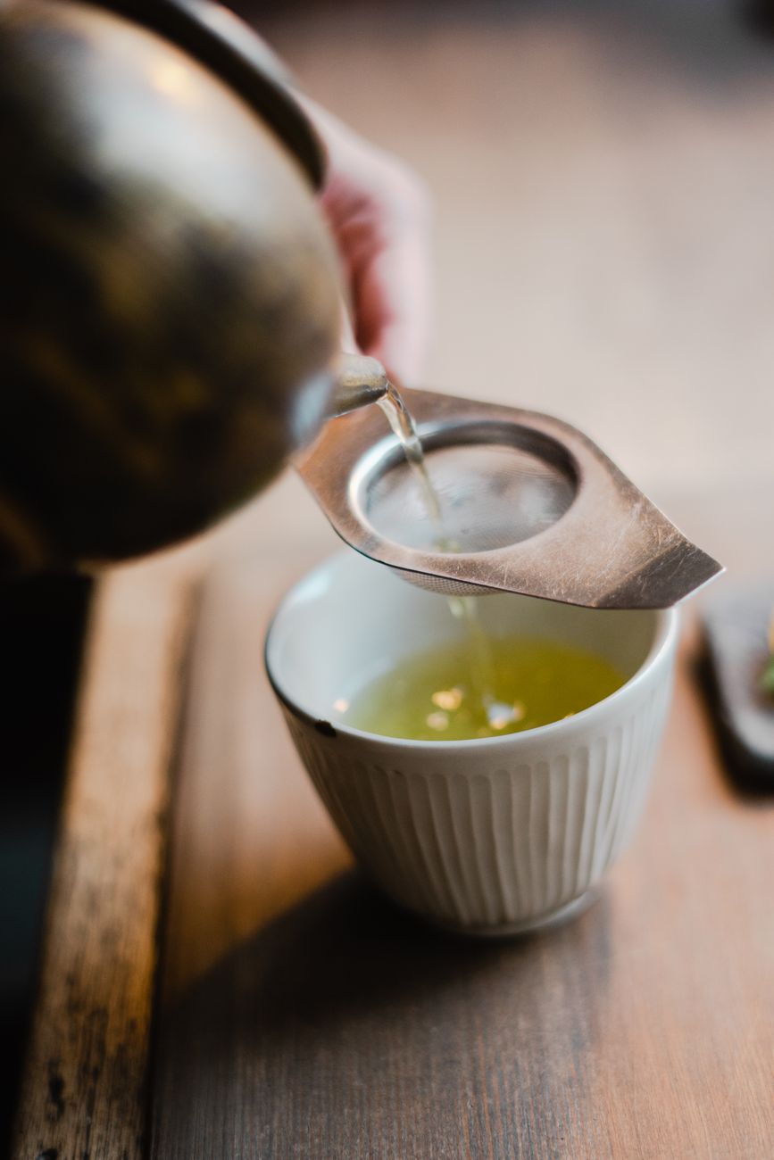 Weathered ceramic tea bowl with natural crack repairs sits on aged wooden surface beside bamboo tea whisk in dim light.