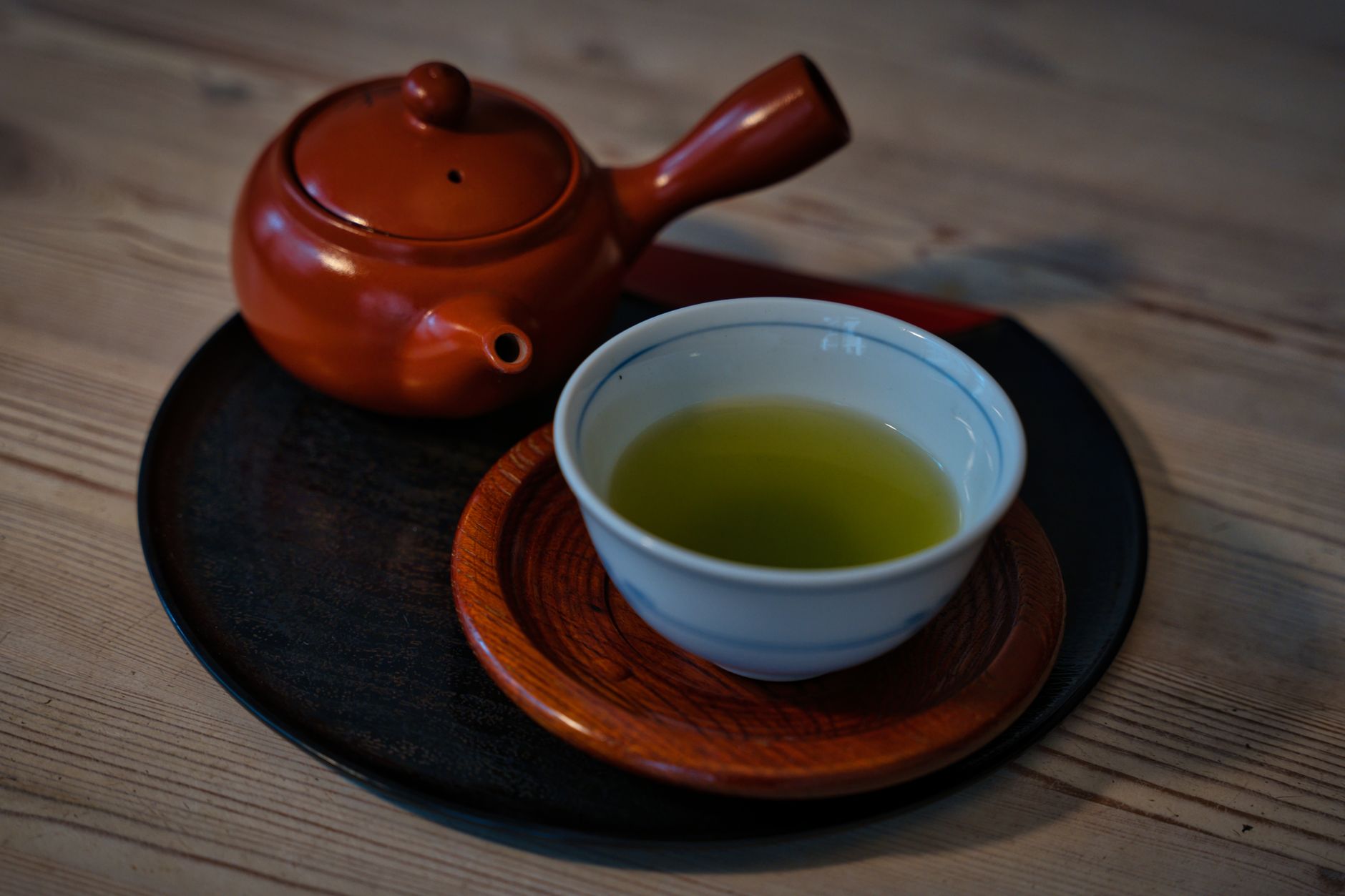 Japanese person pouring green tea from a ceramic kyusu teapot into a small handleless cup on a wooden table.