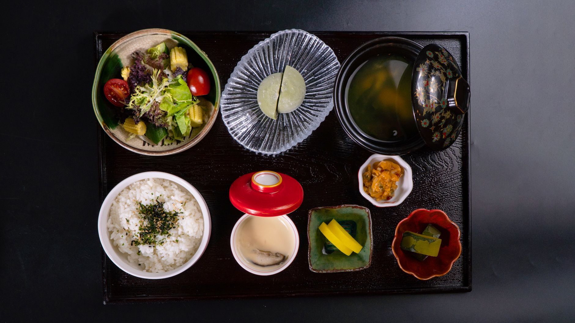 Traditional Japanese meal arrangement showing a bowl of miso soup, rice, grilled fish, pickled vegetables, and simmered vegetables on a wooden tray.