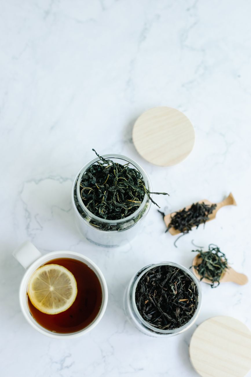 Fresh green tea leaves in a bamboo basket beside dark oxidized black tea leaves on a wooden table.