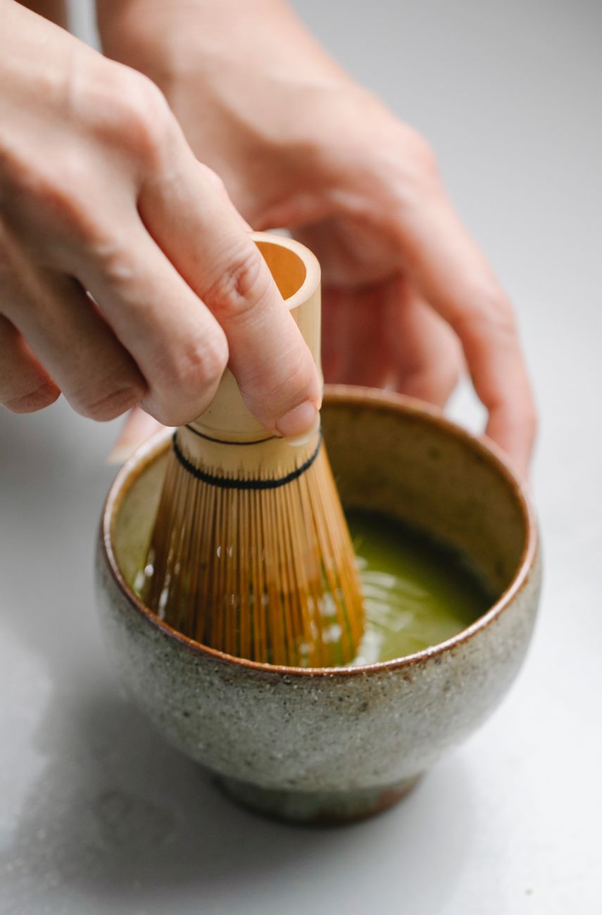 Two wooden scoops holding bright green ceremonial matcha powder beside duller culinary grade matcha on bamboo mat.
